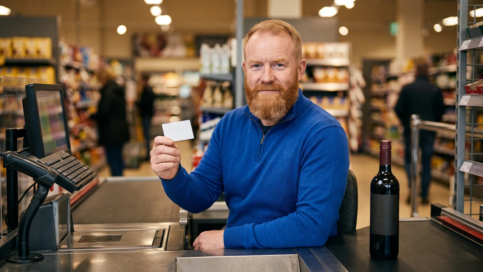 A checkout assistant with a ginger beard sits at his till holding a small plain ID card, with a bottle of wine waiting on the belt beside him.