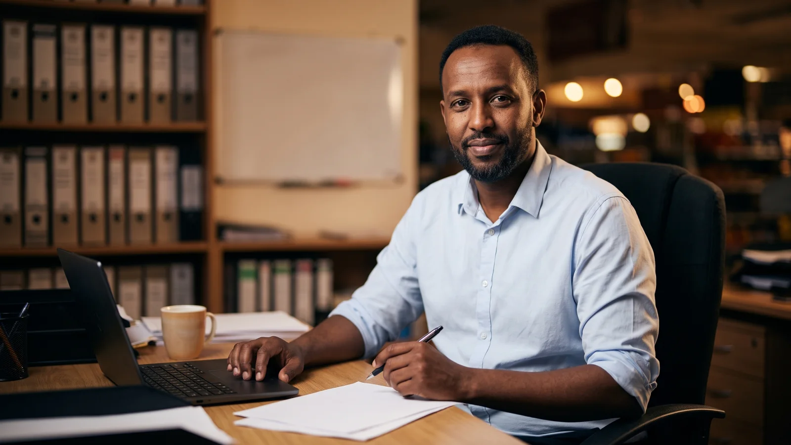 A supermarket manager with a short beard sits at a laptop in a small back office in the afternoon, planning the staff rota.