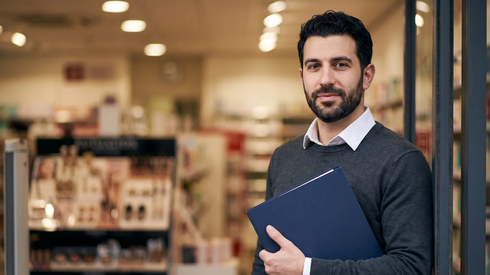 A young man in a plain white shirt and grey jumper holds a closed document folder as he waits for an interview inside a UK pharmacy shop.
