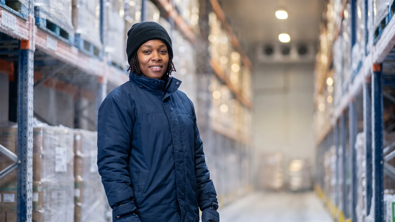 A warehouse operative in a padded freezer suit and hat stands among frosted pallets in a cold store at a regional distribution centre.