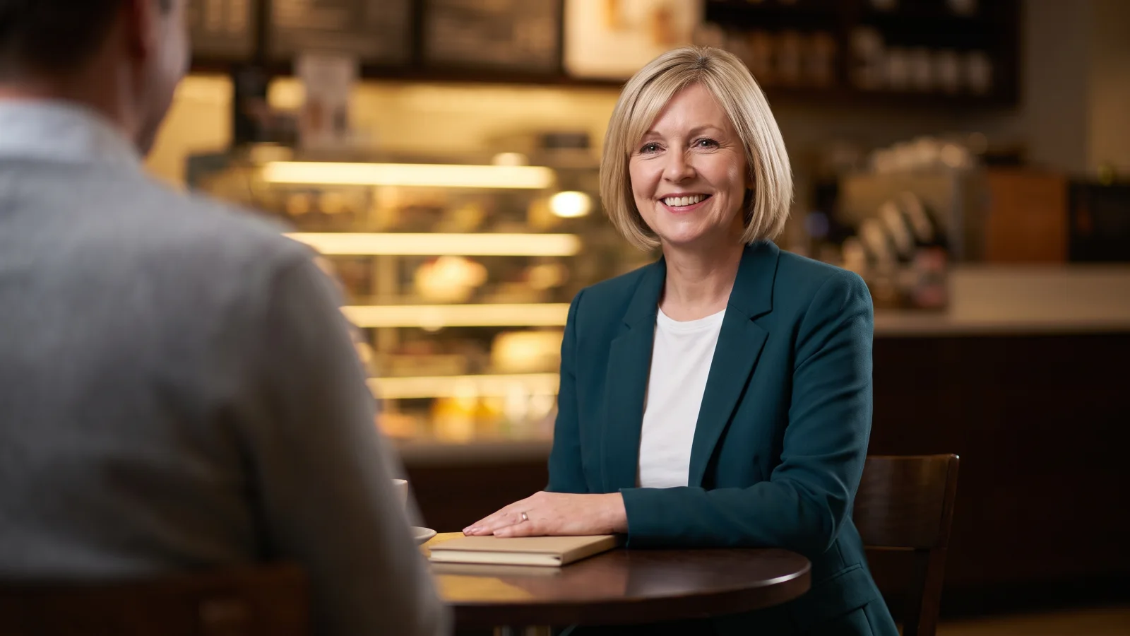A woman in a smart blazer sits at a small café table in a store for her job interview, a hiring manager blurred in the foreground.