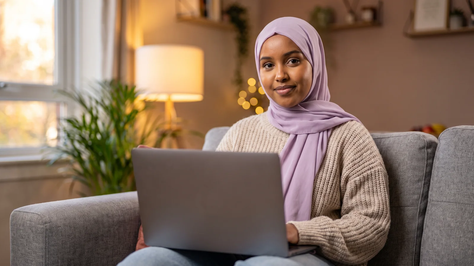 A young woman in a plain headscarf sits on a sofa with a laptop on her knees, ready to start an online job assessment at home.