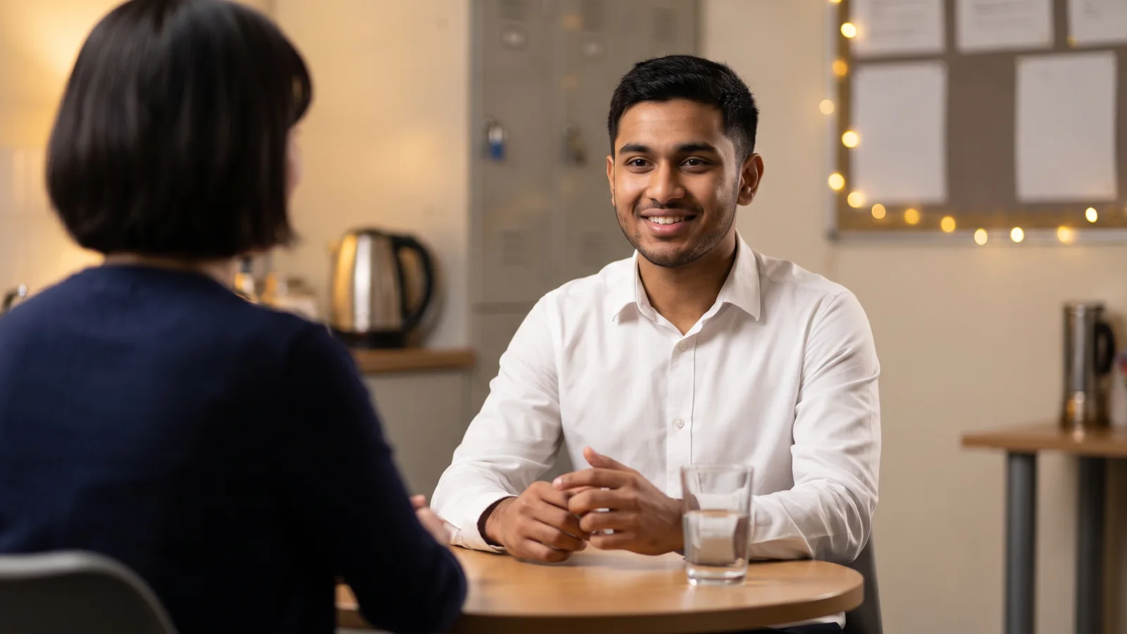 A young man in a plain white shirt sits across a table from an interviewer in a supermarket staff room, smiling as he answers a question.