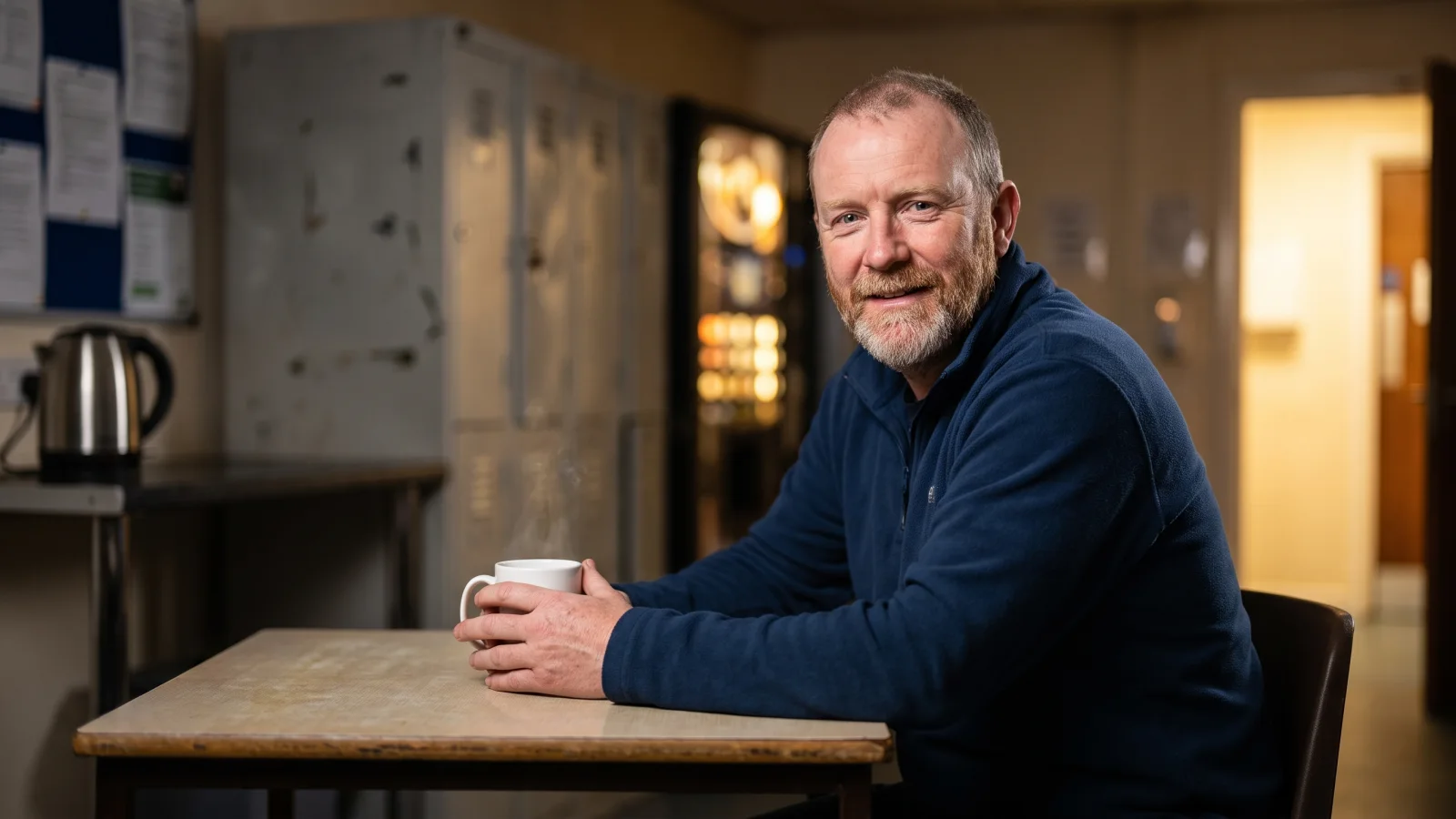 An older night worker with a greying beard sits with a mug of tea on his break in a small, quiet supermarket staff room in the early hours.