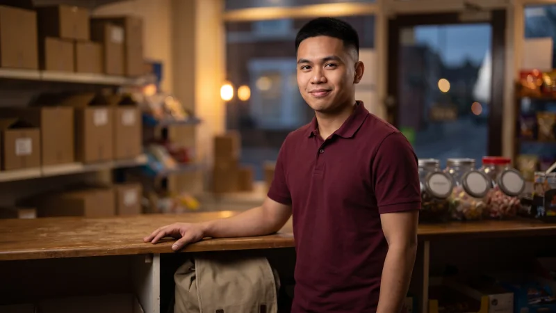 A university student in a plain burgundy polo shirt stands behind the counter of a UK corner shop on a weekday evening, smiling.
