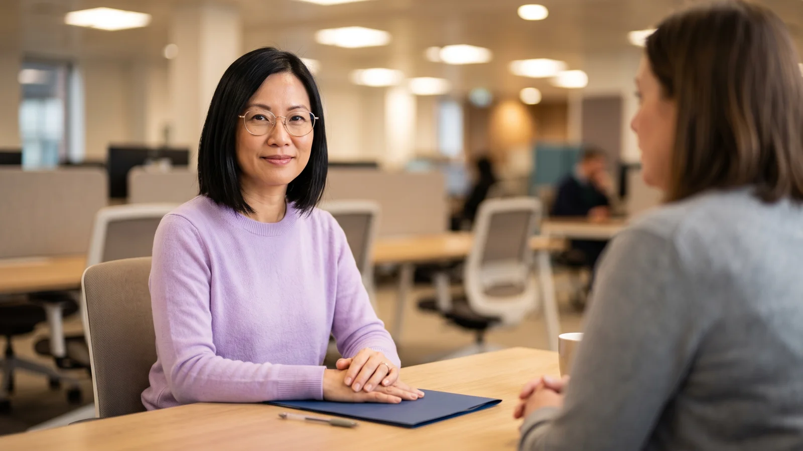 A woman in a plain lilac jumper sits at a desk in a quiet benefits office, a closed folder in front of her, while a work coach sits blurred beside her.