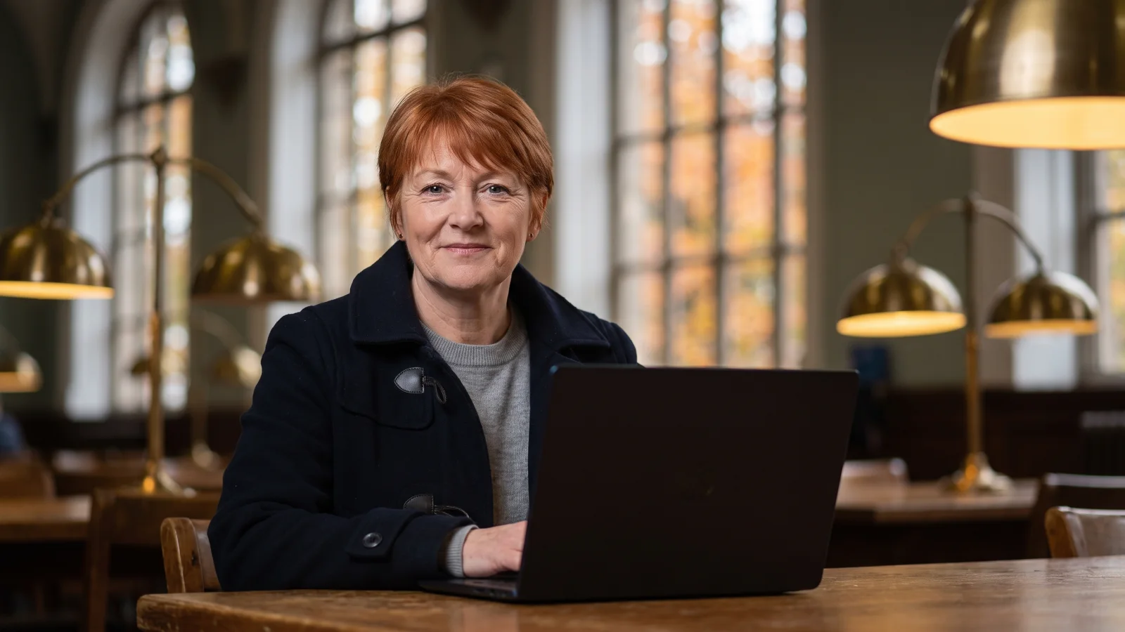 A woman in a navy duffle coat sits at a public library table with a laptop, applying for Christmas shop jobs on an autumn afternoon.