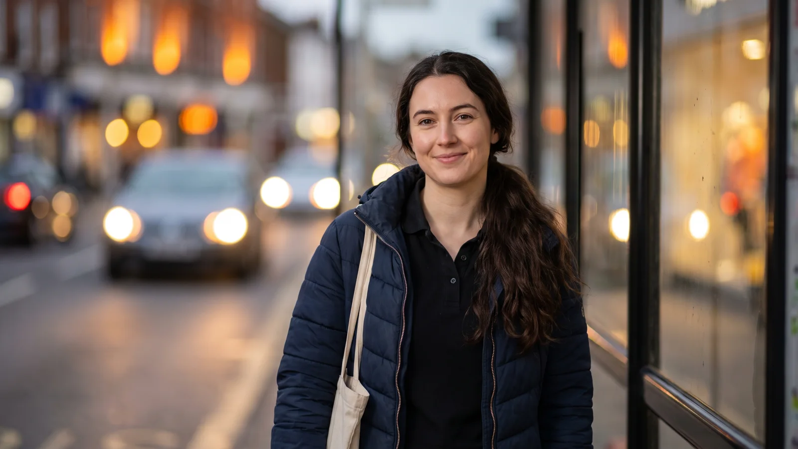 A woman in a plain navy jacket waits at a bus stop on a UK high street in the early evening, heading from one shop job to another.